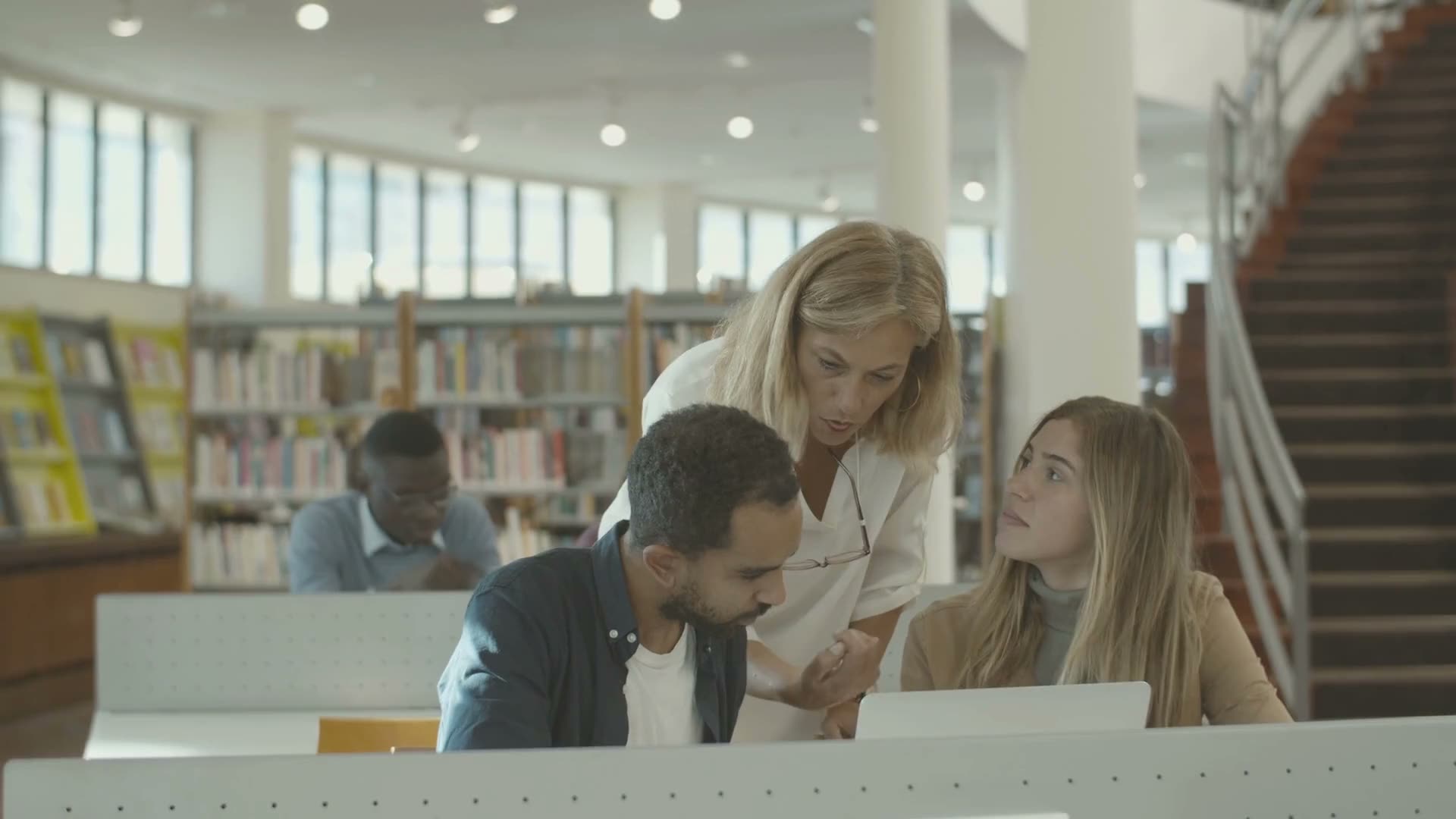 A counselor leans in to help two students at a laptop in a library.