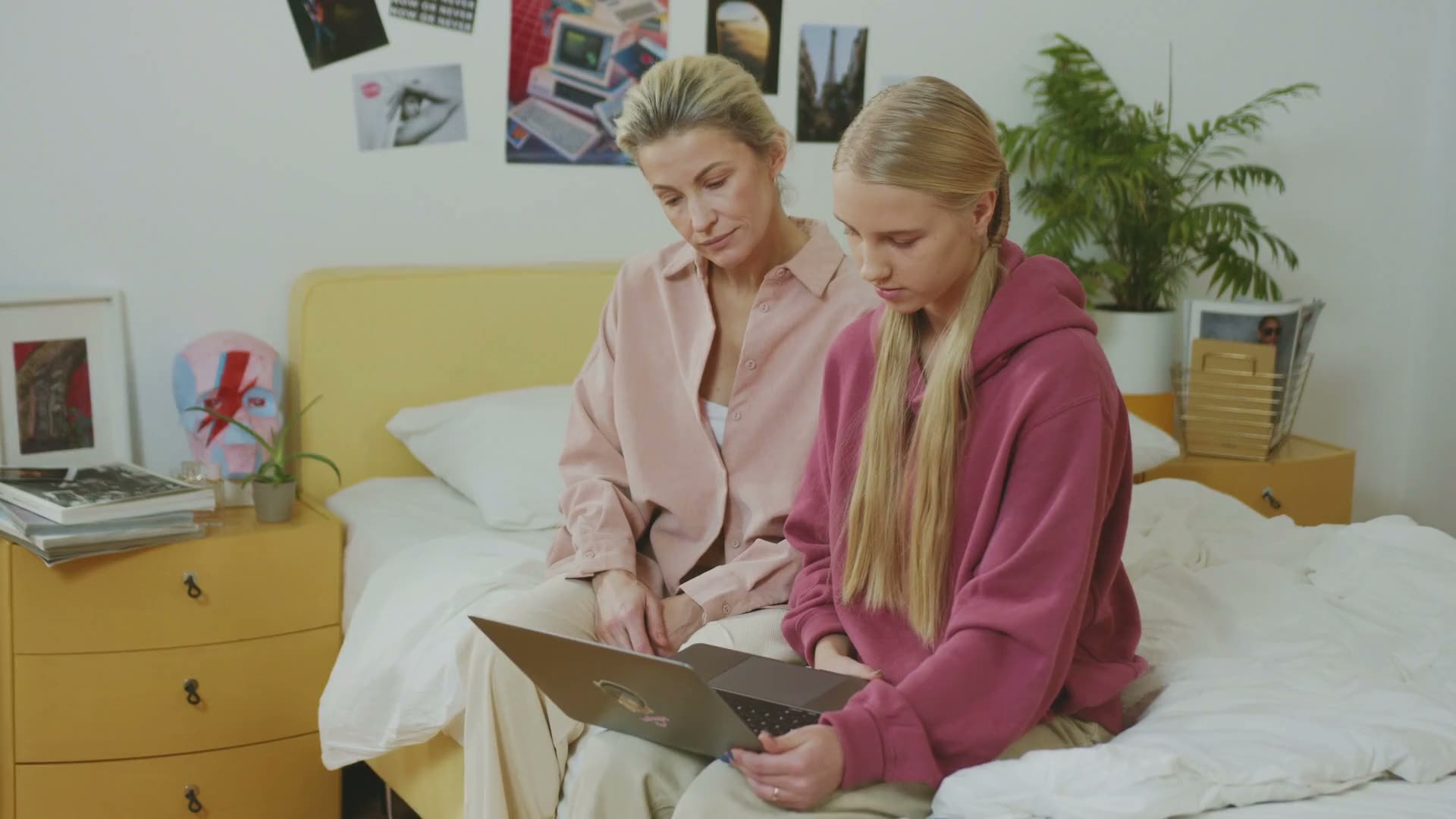 A parent and her daughter sit on a bed and look at a laptop together.
